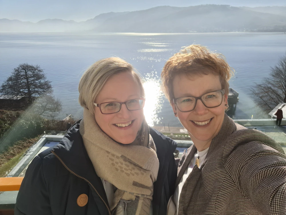 Zwei Frauen lächeln in die Kamera vor einem malerischen See, symbolisieren Achtsamkeit und Kooperation